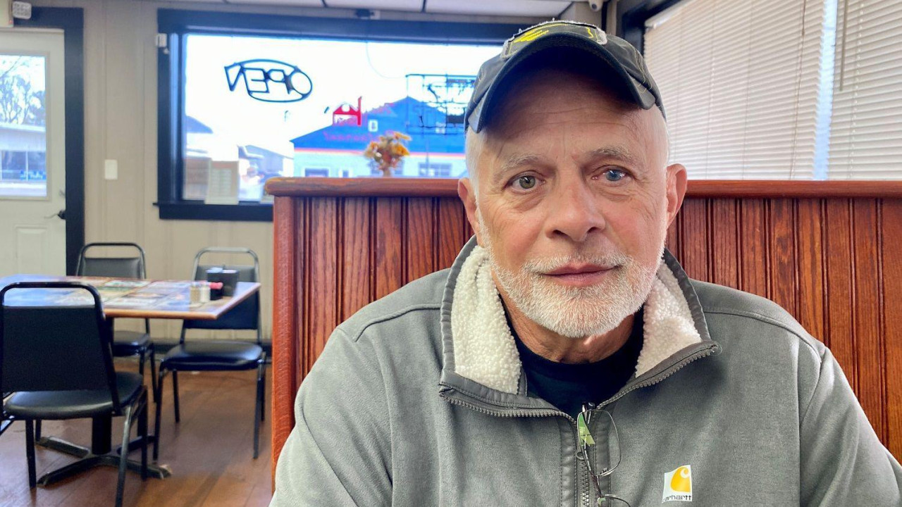 Sedi muškarac sa kačketom i u radnoj odeći sedi u restoranu i gleda pravo u kameru