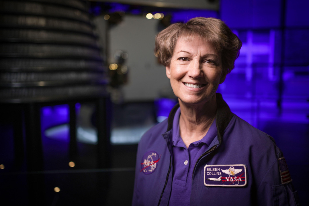 
                    Eileen Collins wearing a blue jacket with a round Nasa patch on one side and a rectangular shuttle patch on the other saying her name. She has short brown hair and brown 