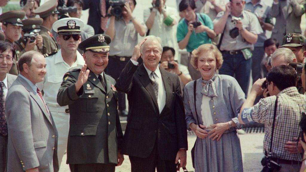 Bivši predsednik SAD Džimi Karter, u crnom odelu sa sivom kravatom i u beloj košulji, sa suprugom Rozalin u sivom kompletu i beloj majici prelazi granicu i ulazi u Severnu Koreju u junu 1994. godine. Oko njih je grupa vojnika i novinari sa fotoaparatima