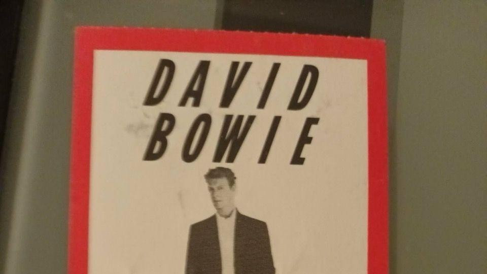 Koncertna ulaznica na kojoj je Dejvid Bouvi u crnim pantalonama i sakou, ispod kojeg ima belu košulju