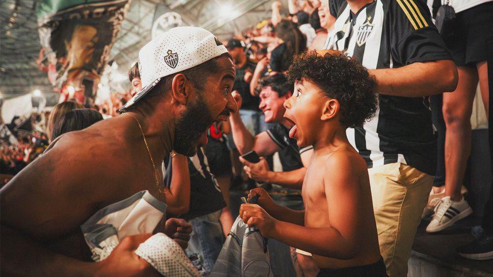 Otac i dete slave gol Atletiko Mineira. Oboje viču jedno na drugo od radosti. Okruženi su navijačima u crno-belim prugastim dresovima.