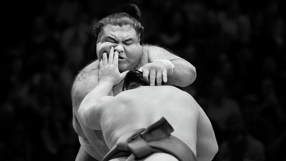 Dva sumo rvača se bore u tradicionalnom ringu, snimljeno u crno-beloj tehnici. Jedan rvač se kreće napred dok se drugi sprema, publika u pozadini je zamućena