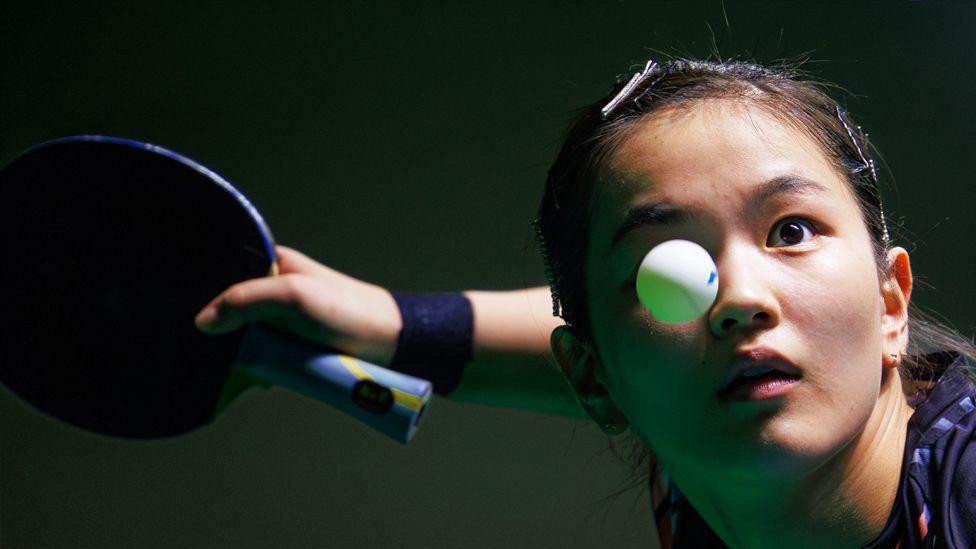 Krupni plan stonoteniserke koja se sprema da reketom udari lopticu, ruka joj je ispružena i na njoj se vidi narukvica. Loptica se vidi u letu iznad desnog oka igrača.