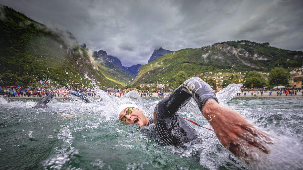 Henri Bramvel-Riks u ronilačkom odelu pliva kroz uzburkanu vodu tokom trke, dok kapljice prskaju okolo. Iza plivača, gomila ljudi se nalazi duž obale, a zelene planine se uzdižu pod oblačnim nebom.
