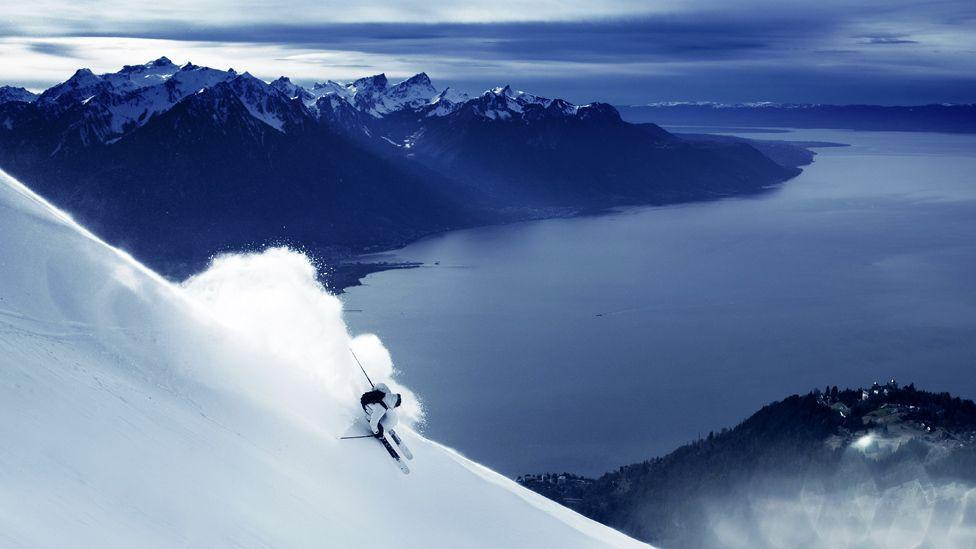 Skijaš se probija kroz svež sneg na strmoj planinskoj padini, stvarajući oblak snežnog praha.
