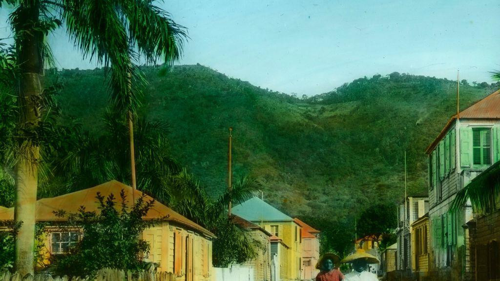 Dve žene sa širokim šeširima i belim suknjama šetaju ulicom sa šarenim kućama u Sent Tomasu na Američkim Devičanskim Ostrvima, na slici iz oko 1890. godine