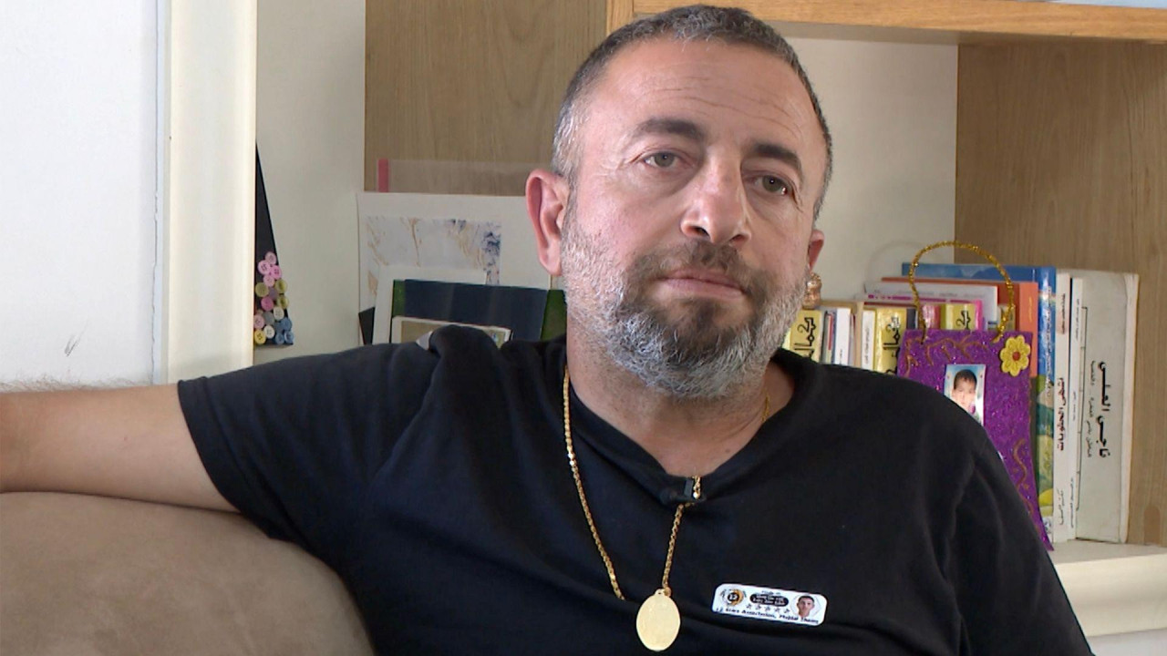 
                    A close-up of a man in a black T-shirt sitting on a sofa. He has short dark hair, a short moustache and a short beard which are all flecked with grey. He is wearing a gol