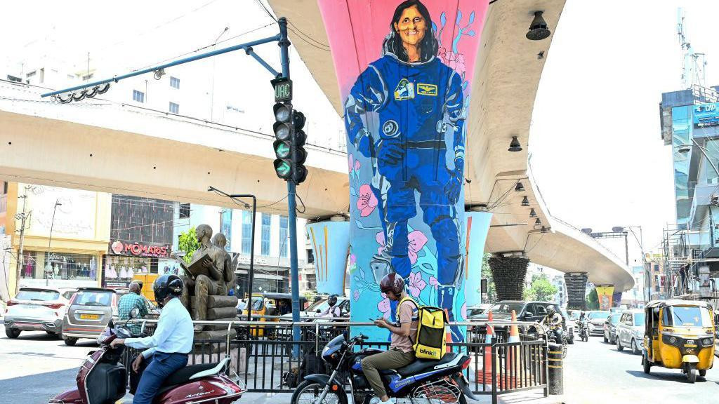 Veliki mural sa likom Sunite Vilijams u plavom svemirskom odelu na stubu jedne od saobraćajnih raskrsnica kuda prolaze motociklisti, automobili i tuk tukovi
