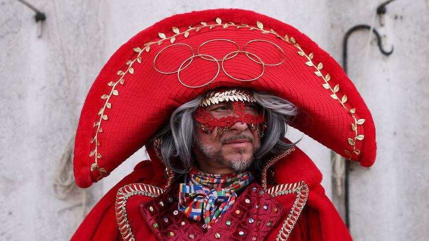 Muškarac na glavi ima crvenu kapu na kojoj zlatnim bojama ušiveni olimpijski krugovi i grančice masline, preko očiju ima crnu maksu