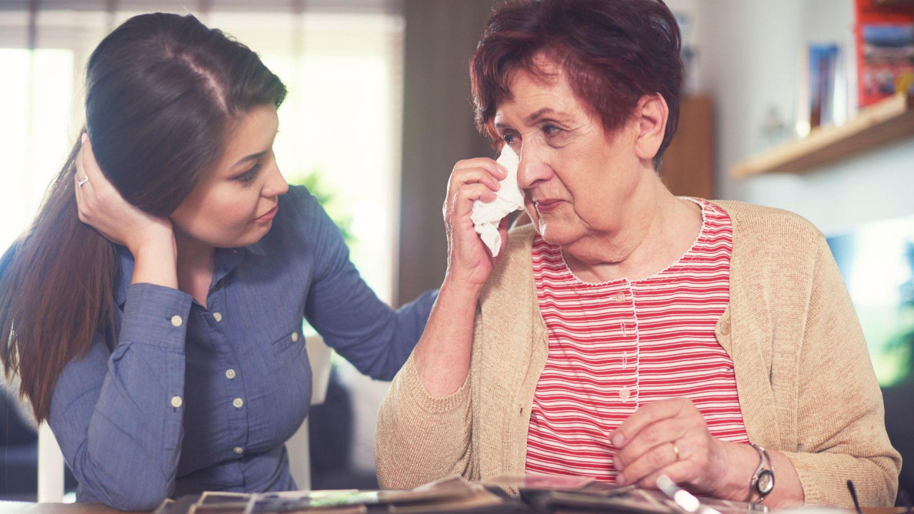 Starija žena drži maramicu na suznom oku dok gleda foto-album; mlađa žena koja sedi pored je teši