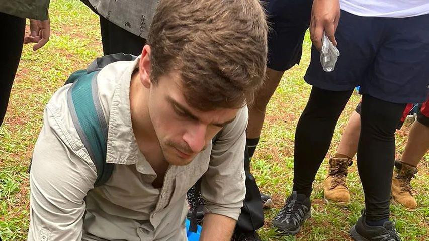 Čovek u svetlo sivoj majici i zelenim pantalonama čuči na travi sa nizom pečuraka poređanih na zemlji ispred njega