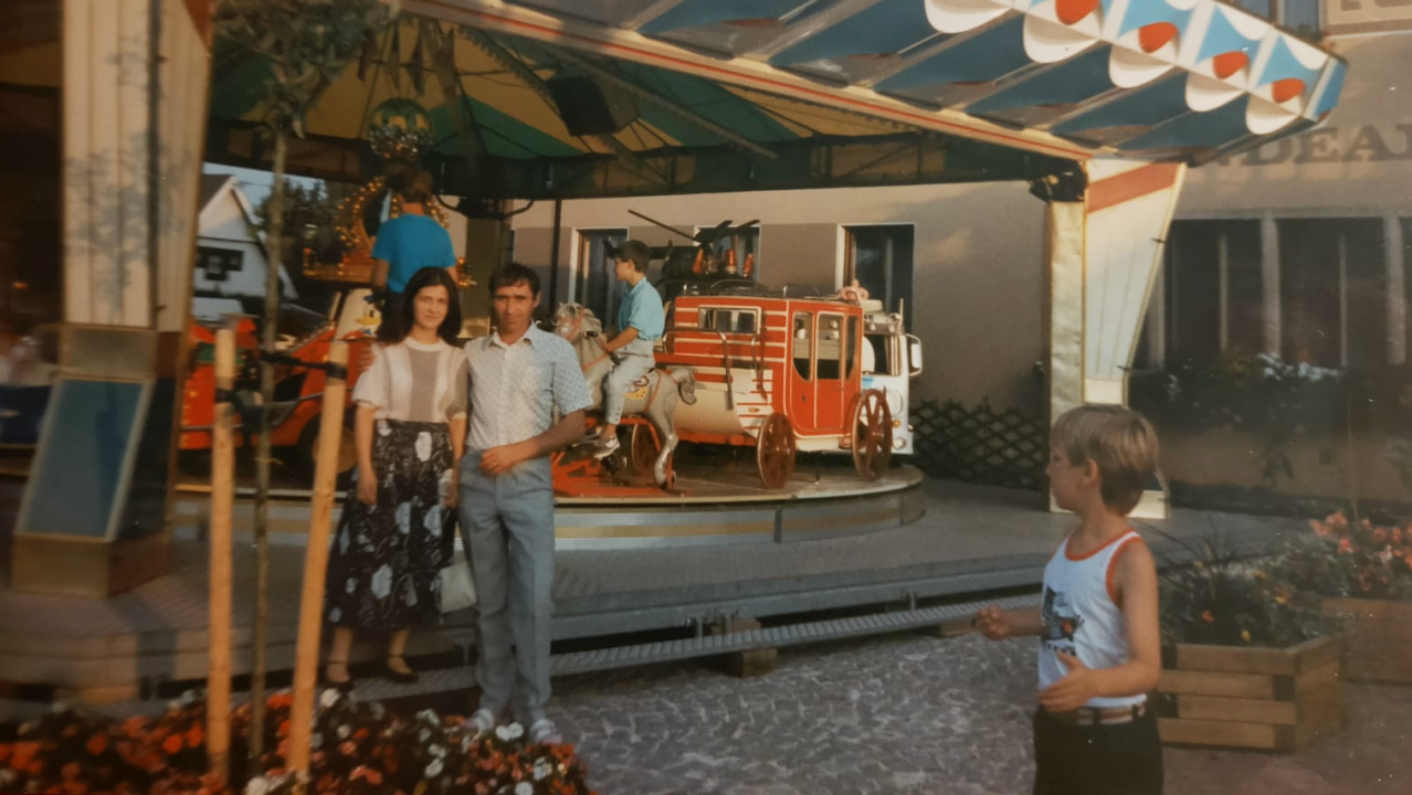 Dvoje ljudi ispred ringišpila i gleda ih plavokosi dečak