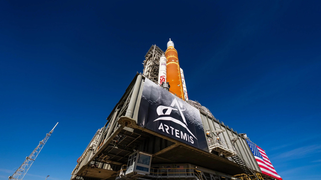 Nasin ogromni sistem za lansiranje u svemir i letelica Orion u svemirskom centru Kenedi na Floridi naspram vedrog plavog neba sa zastavama Artemide i američkom zastavom 