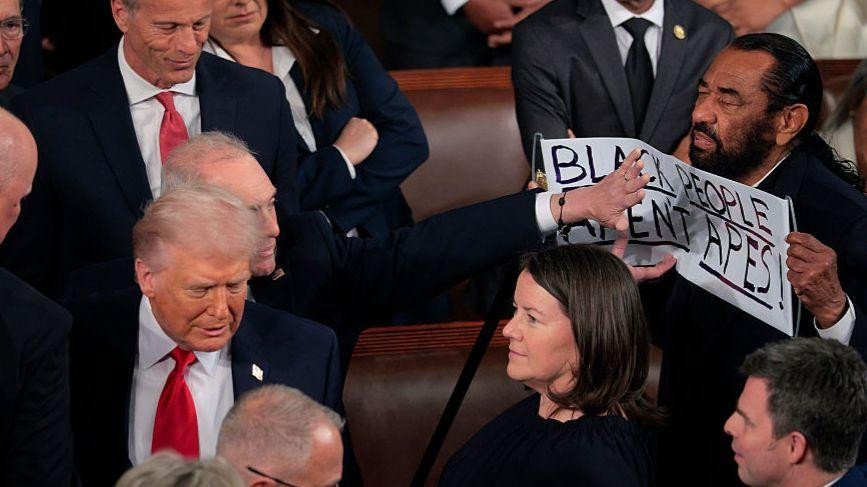Rep Al Green holds up a sign that is being swatted away from fellow lawmakers as Trump passes. It reads, "black people aren't apes".